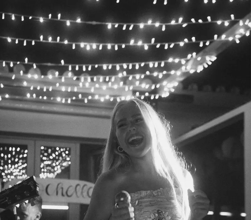 Singer laughing as she performs with a drummer under string lights, black and white photo